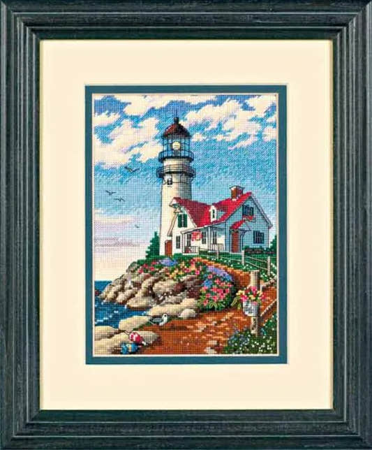Framed cross-stitched picture of a lighthouse on a rocky shore with a blue sky.
