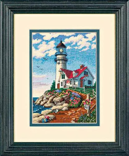 Framed cross-stitched picture of a lighthouse on a rocky shore with a blue sky.