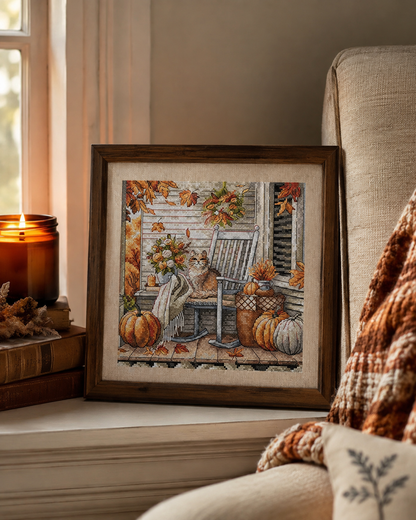 Un ricamo a tema autunnale incorniciato, appoggiato accanto a una sedia in un ambiente accogliente. Il disegno raffigura una scena su un portico con una sedia a dondolo, zucche, foglie autunnali e un cesto, racchiusa in una cornice in legno scuro.
