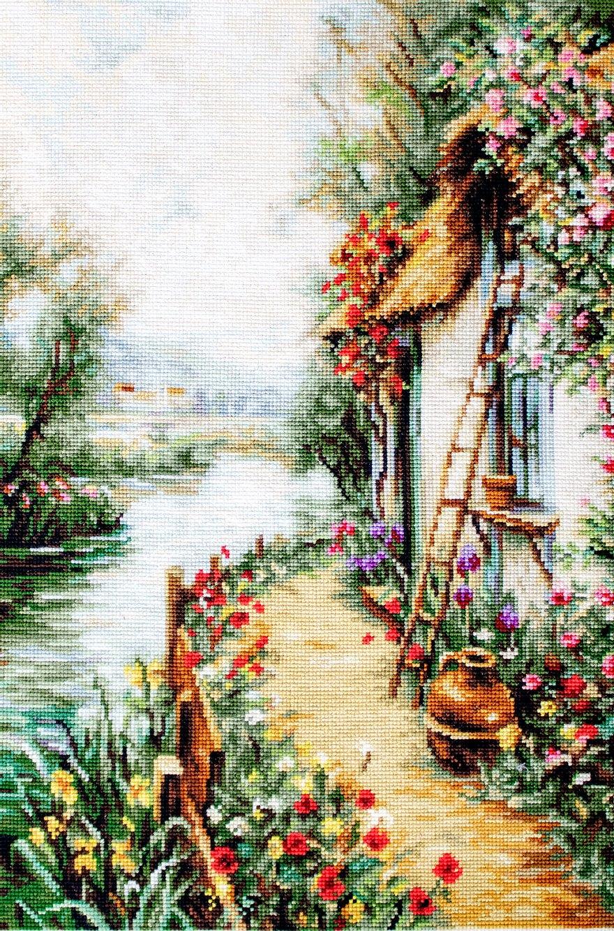 Kit completo da ricamo a mezzo punto Luca-S G581 – Lungo il fiume (Along the River). Paesaggio lungo un fiume con una casa sulla riva, fiori rossi e vegetazione rigogliosa.